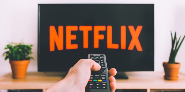 A TV with the Netflix logo sits behind a hand holding a remote