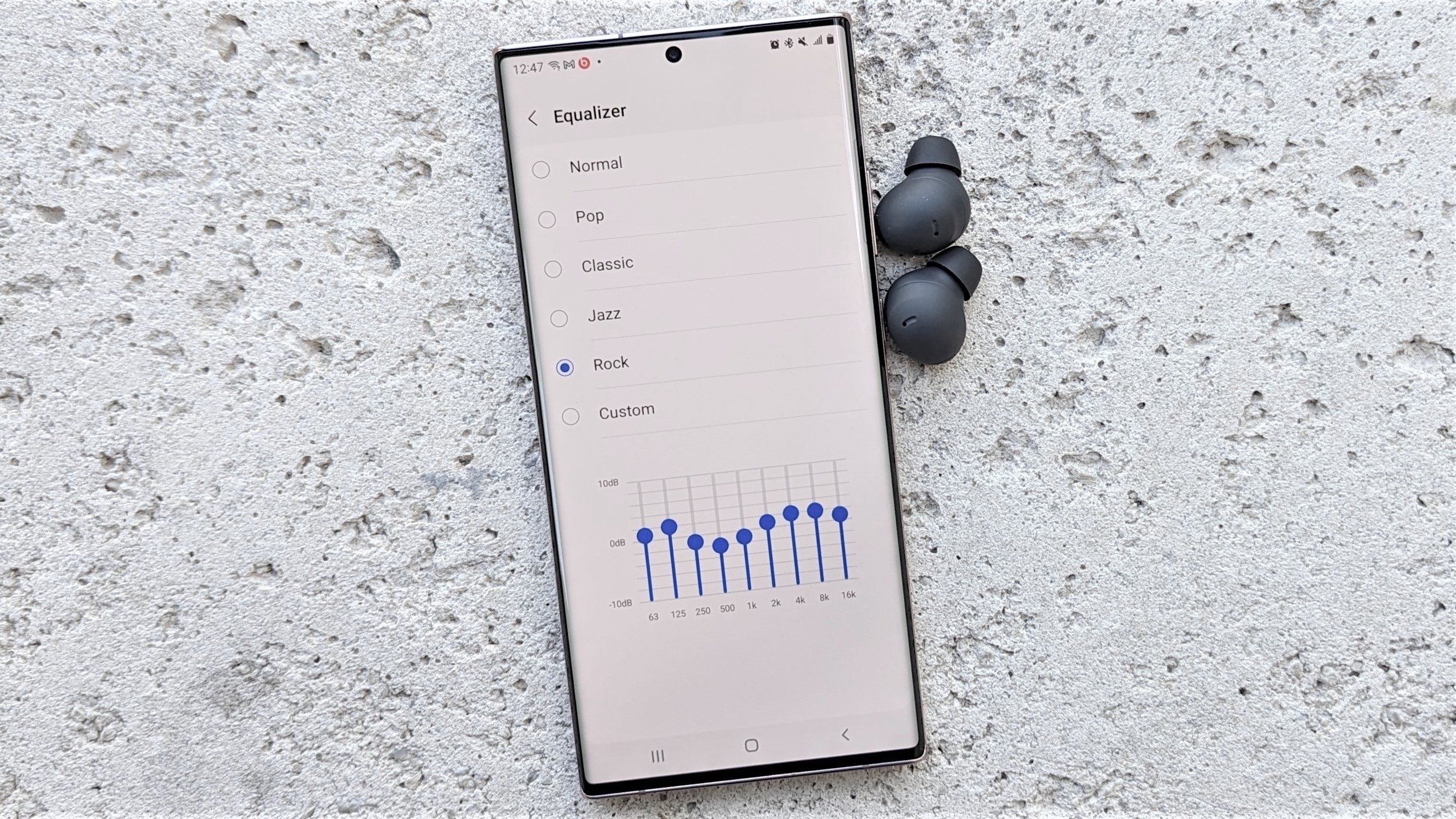 هاتف Samsung Galaxy الذكي المزود بسماعات أذن موضوعة على سطح حجري تعرض إعدادات تخصيص EQ